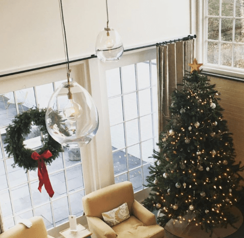 A cozy room with a Christmas tree, decorated with lights and ornaments, and a wreath hanging by large windows.