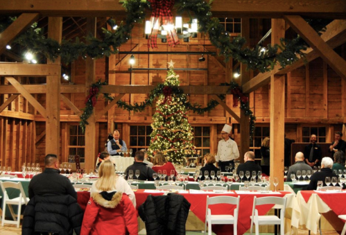 A festive dining setup in a rustic barn, featuring a Christmas tree and guests preparing for a holiday meal.