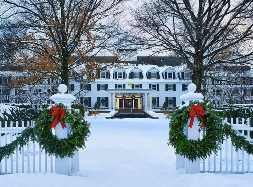 A snowy scene of a grand white hotel, adorned with festive wreaths, surrounded by bare trees.