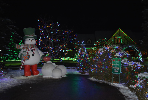 A large snowman with a top hat stands beside decorated trees and colorful lights in a winter night scene.