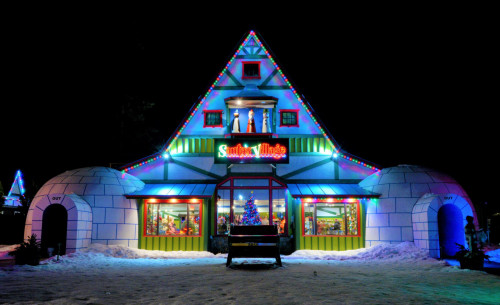 A colorful, festive building with Christmas lights, resembling a house and igloos, set in a snowy landscape at night.