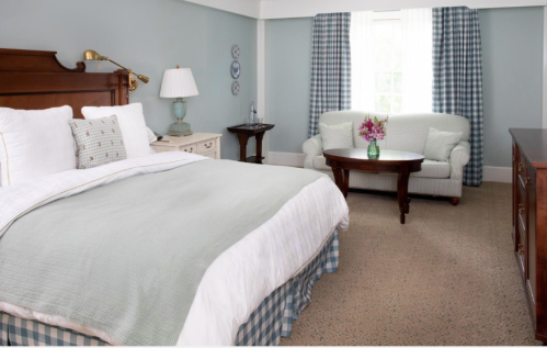Cozy bedroom with a large bed, a sofa, a coffee table, and soft blue walls, featuring natural light from a window.