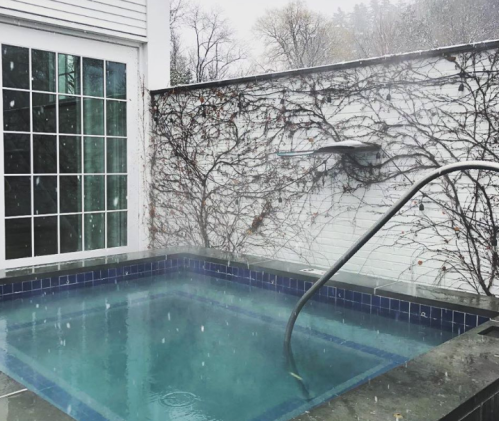A serene hot tub surrounded by ivy-covered walls, with snow gently falling in a tranquil winter setting.