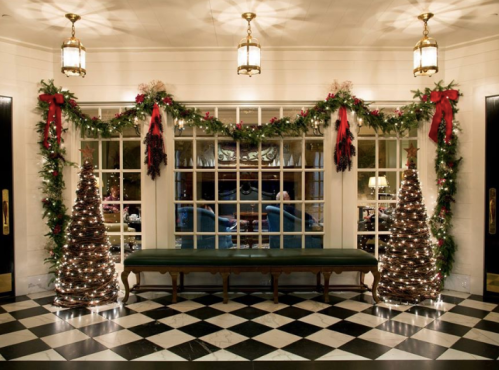 A festive entryway decorated with garlands, lights, and two Christmas trees, featuring a checkered floor and elegant lighting.