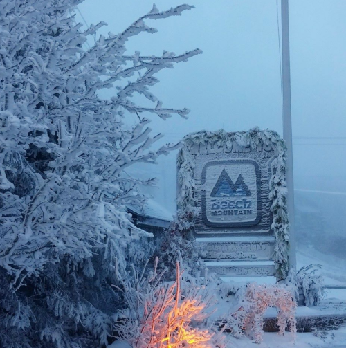 A snowy scene featuring a sign for Beech Mountain, surrounded by frosted trees and a soft orange glow.