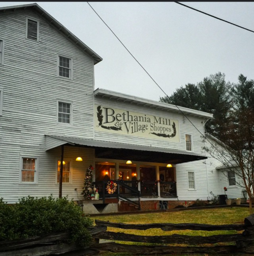 Historic white building with "Bethania Mill & Village Shoppes" sign, surrounded by greenery and festive decorations.