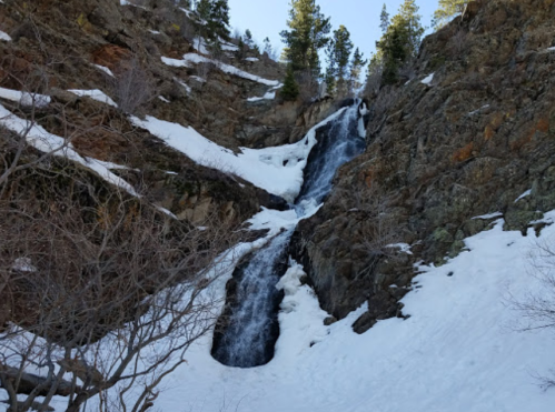 A waterfall cascading down rocky terrain, surrounded by snow and sparse trees under a clear blue sky.