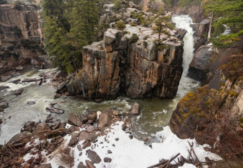 A rocky landscape with a waterfall cascading into a river, surrounded by trees and patches of snow.