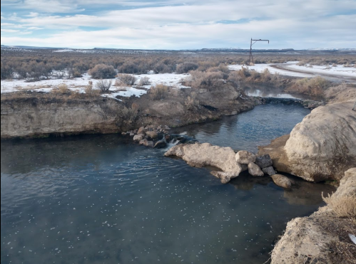 A serene landscape featuring a winding stream surrounded by dry brush and snow, with a distant road and utility pole.
