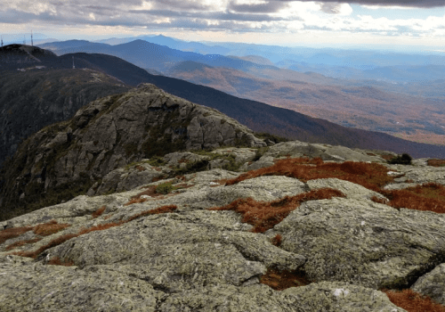 A rocky mountain peak overlooks a vast landscape of rolling hills and valleys, with autumn foliage in the distance.