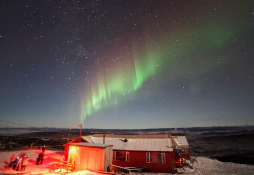 A cabin under a starry sky with vibrant green auroras illuminating the night. Snow-covered landscape in the background.