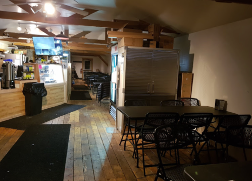 Interior of a casual dining space with wooden floors, tables, and a kitchen area in the background.