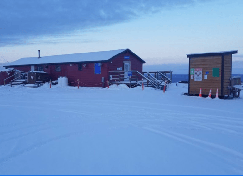 A red building with a sloped roof surrounded by snow, with a wooden sign and cones in the foreground.
