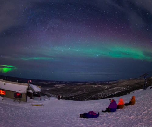 People sitting on snow, gazing at a starry sky with vibrant auroras over a mountainous landscape.