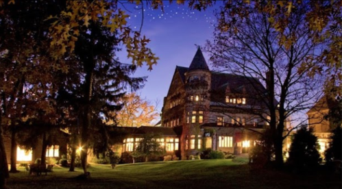 A grand, historic mansion illuminated at dusk, surrounded by trees and a serene lawn.