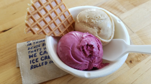 A cup of ice cream with two scoops: one purple and one beige, accompanied by a waffle cone and a napkin.