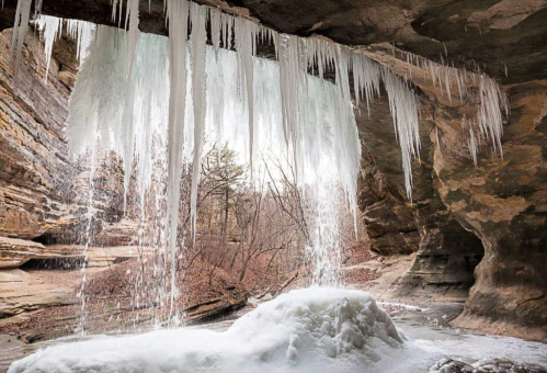 Icicles hang from a rocky overhang, with a frozen waterfall and a snowy landscape in a serene, winter setting.