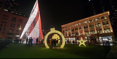 A festive scene at night featuring a large illuminated Christmas tree, ornaments, and people enjoying the holiday decorations.