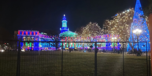 Colorful holiday lights illuminate a building and trees at night, with a festive atmosphere and a fenced area in the foreground.