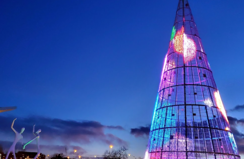 A brightly lit, colorful Christmas tree stands against a twilight sky, with two abstract figures dancing nearby.