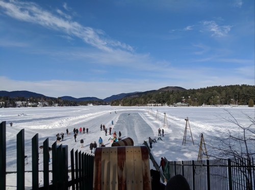 A snowy landscape with people skating on a frozen lake, surrounded by mountains and trees under a clear blue sky.