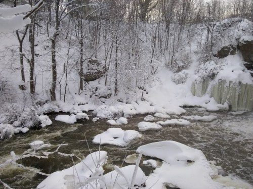 A winter scene of a river with ice and snow, surrounded by trees and a frozen waterfall in the background.
