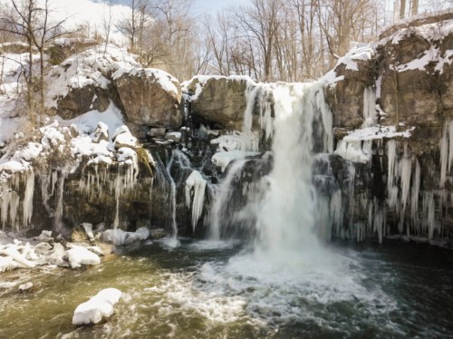 A frozen waterfall surrounded by snow-covered rocks and trees, with icicles hanging from the cliff edges.