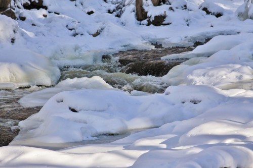 A snowy landscape with a flowing river partially covered in ice and snow, surrounded by trees.