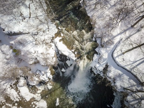 Aerial view of a snowy landscape with a waterfall cascading into a river, surrounded by bare trees.
