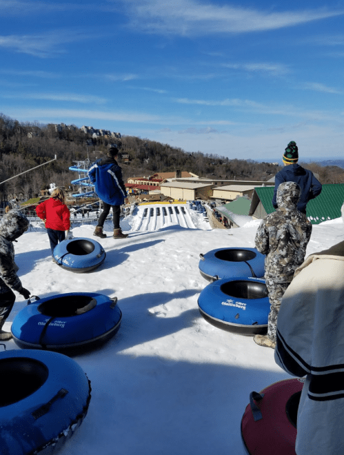 A group of people in winter attire stand at the top of a snow-covered hill, ready to go tubing down the slope.