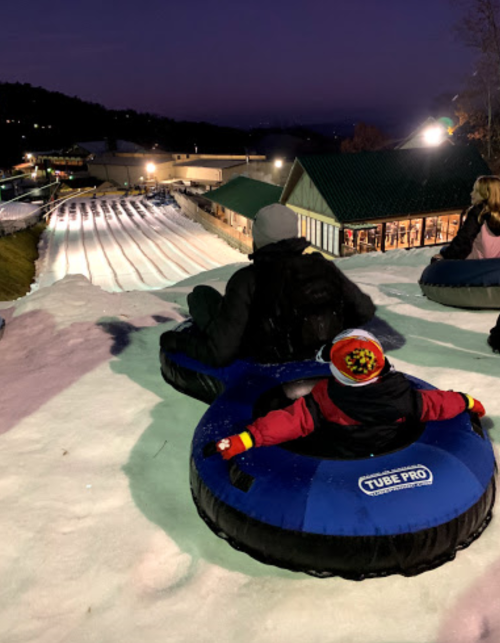 A person and a child sit in snow tubes at the top of a snowy hill, ready to slide down a tubing slope at night.
