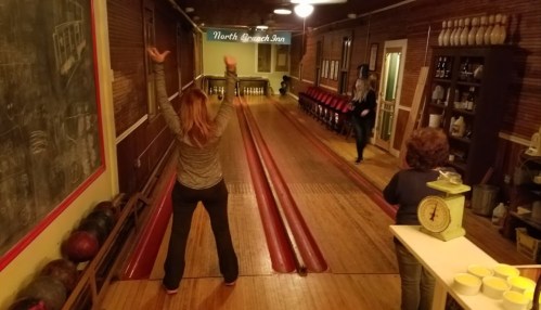 A vintage bowling alley with two women playing, one in motion and another celebrating, surrounded by retro decor.