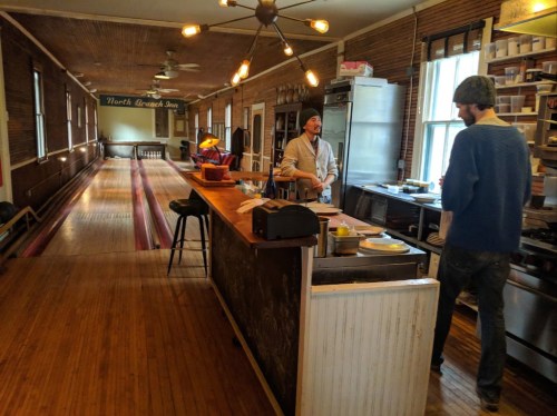 Two people converse in a rustic kitchen with wooden floors and a bowling lane in the background at North Branch Inn.