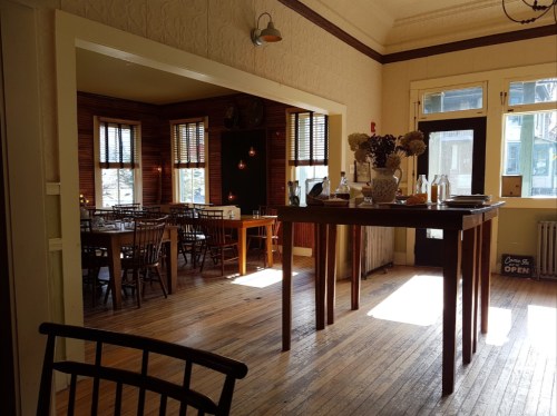 Cozy restaurant interior with wooden floors, tables, and warm lighting, featuring a large table in the foreground.