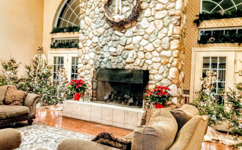 Cozy living room with a stone fireplace, decorated Christmas trees, and festive plants. Warm and inviting atmosphere.