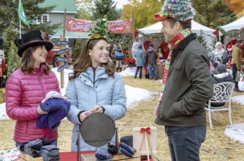 Three people in winter attire smile and chat at a festive outdoor market, surrounded by holiday decorations and snow.