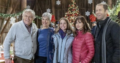 A group of five people smiles together in a festive barn decorated for the holidays, with a Christmas tree in the background.