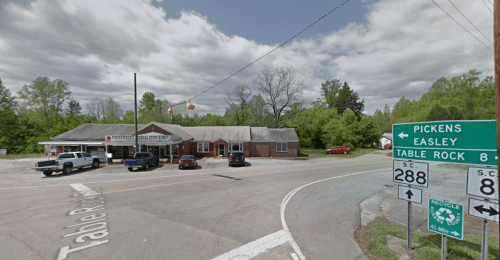 A roadside view of a small building with cars parked outside, near a sign for Table Rock and directions to Pickens and Easley.
