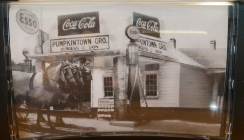 Historic black-and-white photo of a gas station with a horse-drawn carriage and vintage Coca-Cola signage.