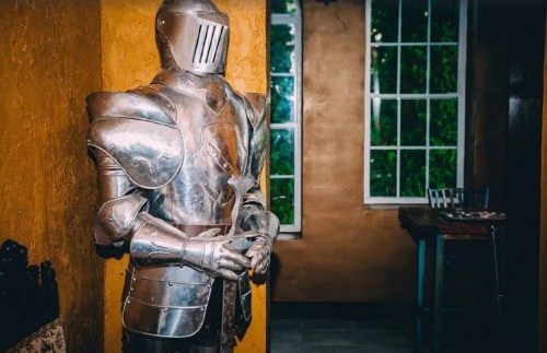 A suit of medieval armor stands against a yellow wall, with windows and a chair visible in the background.