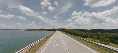 A road stretches over a lake, flanked by greenery and under a partly cloudy sky.