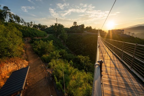 A scenic view of a suspension bridge at sunset, surrounded by lush greenery and a winding path below.