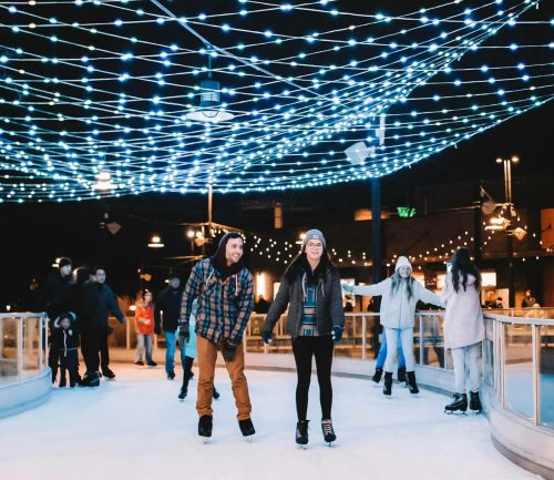 A couple ice skating under a canopy of blue lights, surrounded by other skaters in a festive atmosphere.