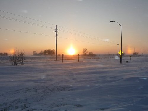 A snowy landscape at sunset, with power lines and a distant road sign silhouetted against the colorful sky.