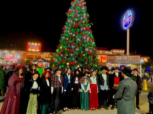 A group of carolers in festive attire stands in front of a brightly lit Christmas tree at night.