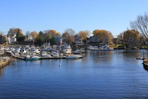 A serene marina with boats docked, surrounded by trees with autumn foliage under a clear blue sky.