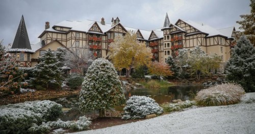 A picturesque hotel surrounded by snow-covered trees and a serene pond, set against a cloudy winter sky.