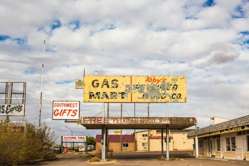 Abandoned gas station with faded signage and overgrown vegetation under a cloudy sky.
