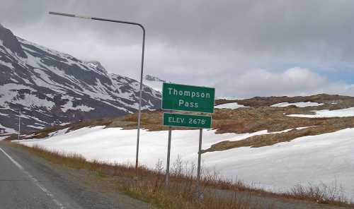 Sign for Thompson Pass with elevation 2678 feet, surrounded by snow-covered mountains and cloudy skies.