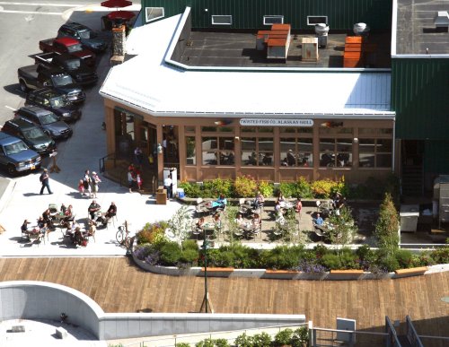 Aerial view of a restaurant with outdoor seating, surrounded by greenery and parked cars. People are dining outside.